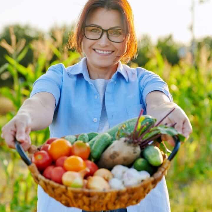 A woman stands in a garden holding a basket filled with fresh vegetables including tomatoes, cucumbers, mushrooms, and beets.