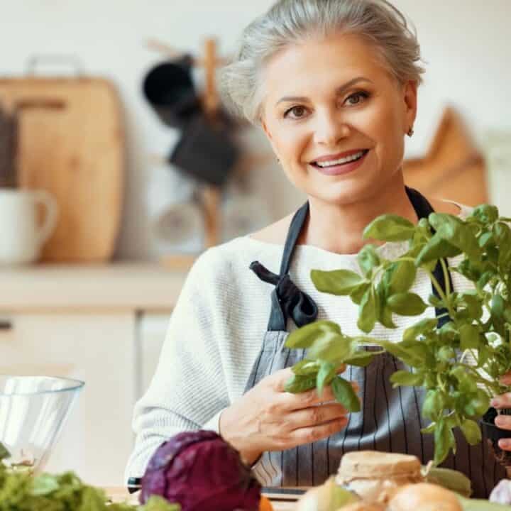 Older woman with gray hair holds fresh basil in a kitchen, surrounded by vegetables and ingredients, smiling at the camera.