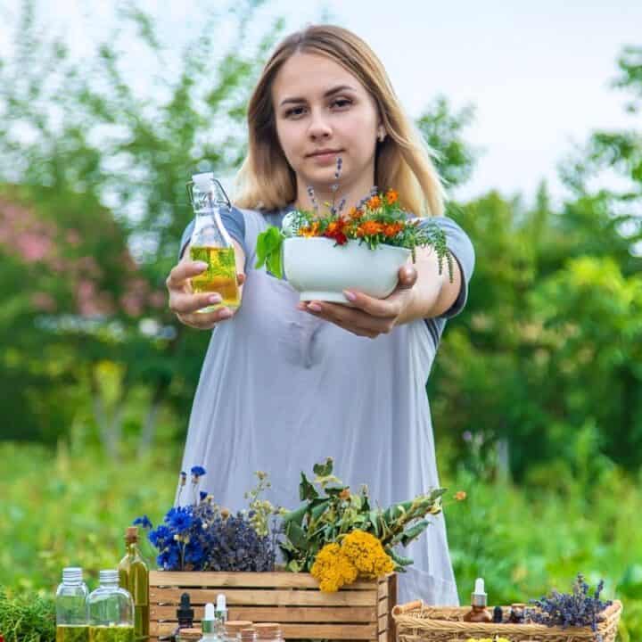 A woman stands outdoors holding a white bowl of flowers and a bottle of oil, with a table of herbs, flowers, and bottles in front of her.