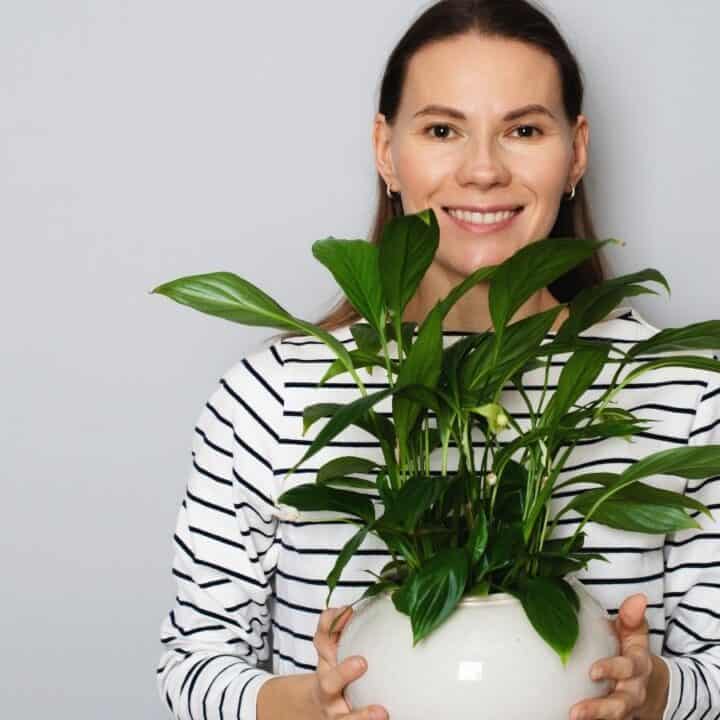 A woman in a striped shirt smiles while holding a white pot with a leafy green plant in front of a plain light gray background.
