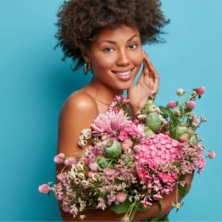 A woman with curly hair smiles and holds a large bouquet of pink and purple flowers against a solid blue background.