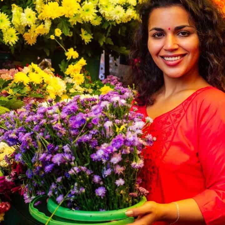 A woman in a red top stands among colorful flowers, smiling and holding a green basket filled with purple blooms.