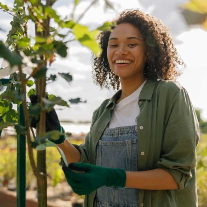 A person wearing green gloves and overalls tends to a plant outdoors in a garden, smiling at the camera on a sunny day.