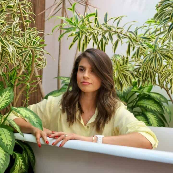 A woman in a light yellow shirt sits in a white bathtub surrounded by various green leafy plants.