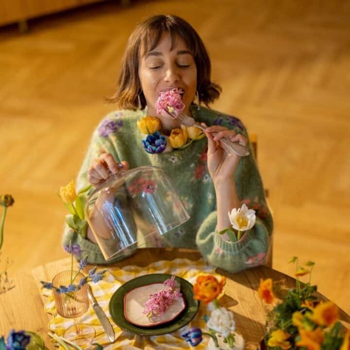 A woman sits at a table decorated with flowers, holding a glass cloche and about to eat a dessert from a fork with her eyes closed.