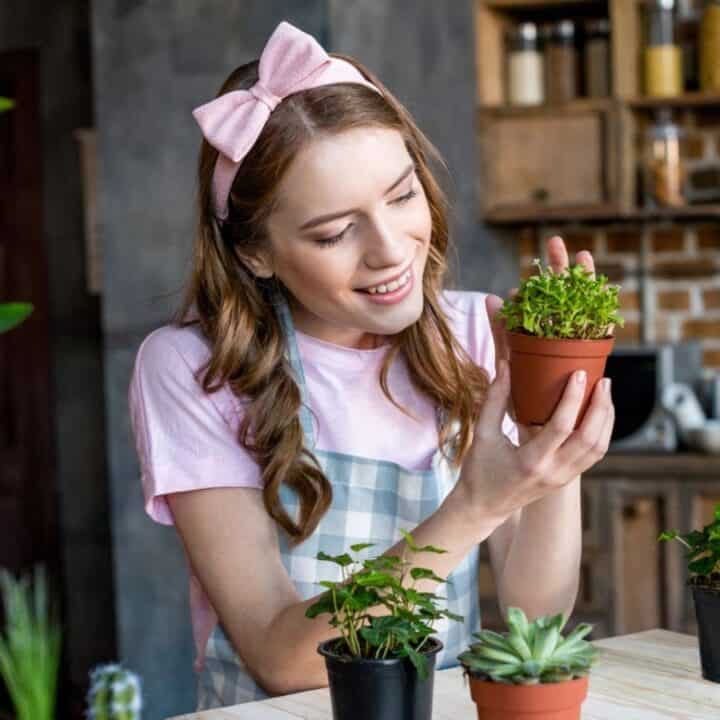A woman wearing a pink headband smiles while holding a small potted plant, surrounded by other plants on a table in a kitchen.