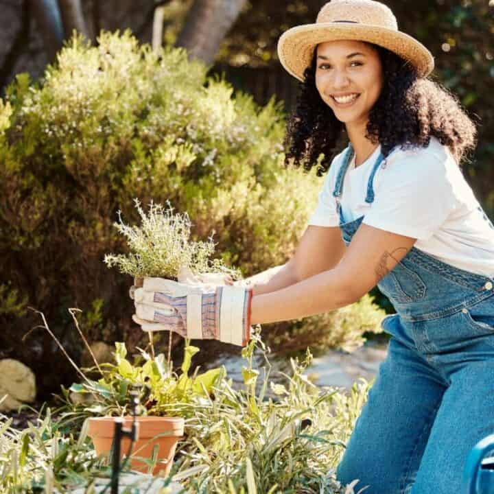 A woman wearing a hat and overalls kneels in a garden, holding a potted plant and smiling at the camera, surrounded by greenery and gardening tools.