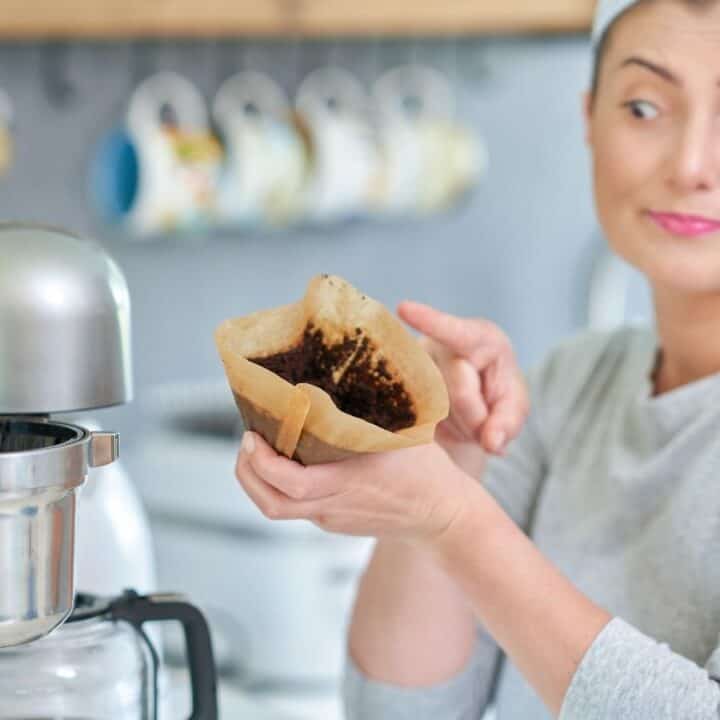 A woman holds a used coffee filter with grounds and points at it, standing next to a coffee maker in a kitchen.