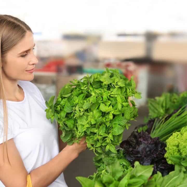 A woman in a white shirt holds a bunch of fresh leafy greens while shopping at a market produce stand.