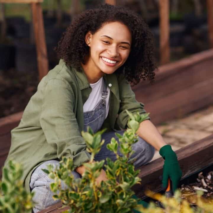 A person wearing a green shirt and gloves smiles while gardening, surrounded by green and yellow plants in a raised garden bed.