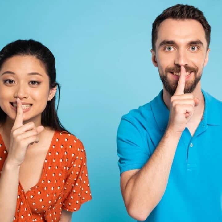 A woman and a man stand side by side against a blue background, each holding a finger to their lips in a shushing gesture.