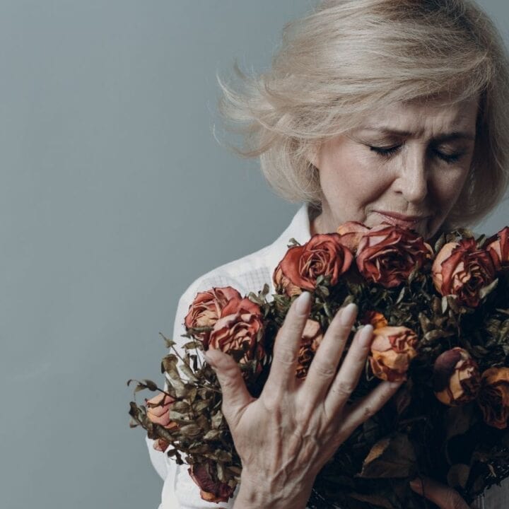 Older woman with short blonde hair holding and looking at a wilted bouquet of dried roses against a plain gray background.