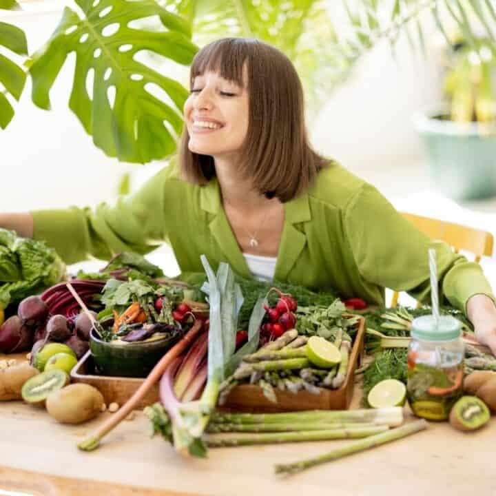 A woman in a green shirt sits at a table covered with assorted fresh vegetables and fruits, smiling with eyes closed.