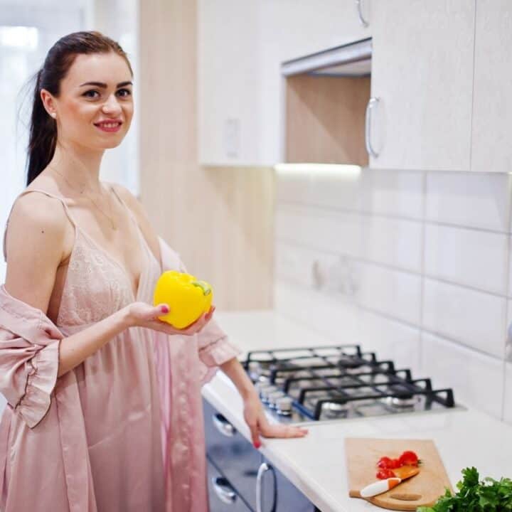 Woman in a pink robe stands in a kitchen holding a yellow bell pepper, with chopped vegetables and herbs on the counter beside her.