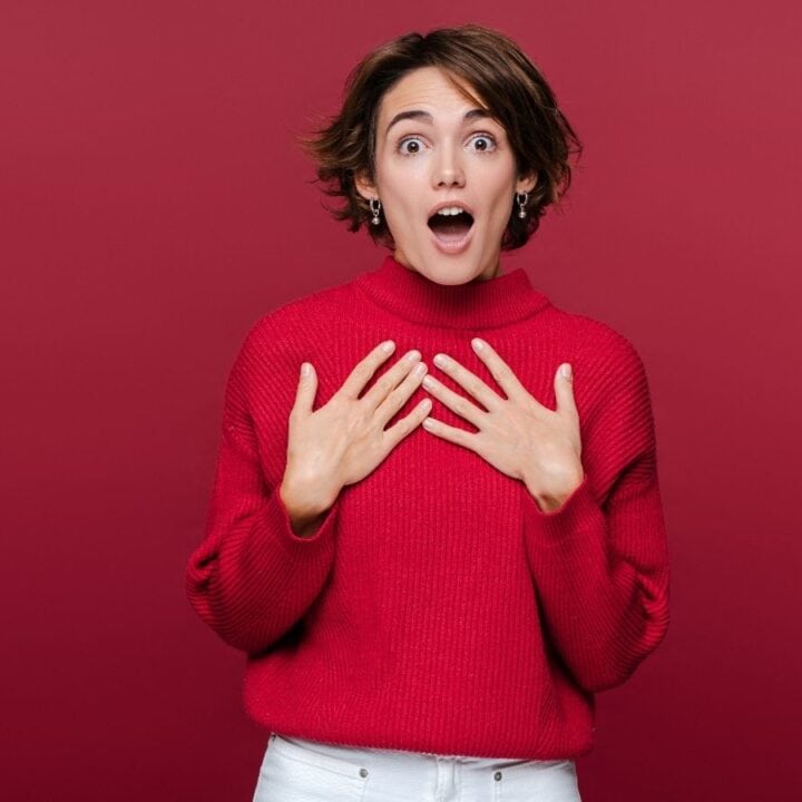 A person wearing a red sweater and white pants stands against a red background, looking surprised with hands held to their chest and mouth open.