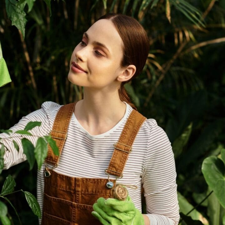 A woman wearing brown overalls and green gardening gloves tends to plants in a lush, green garden.