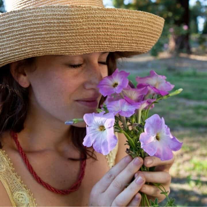 A woman wearing a straw hat and red necklace holds and smells a bunch of purple flowers outdoors on a sunny day.