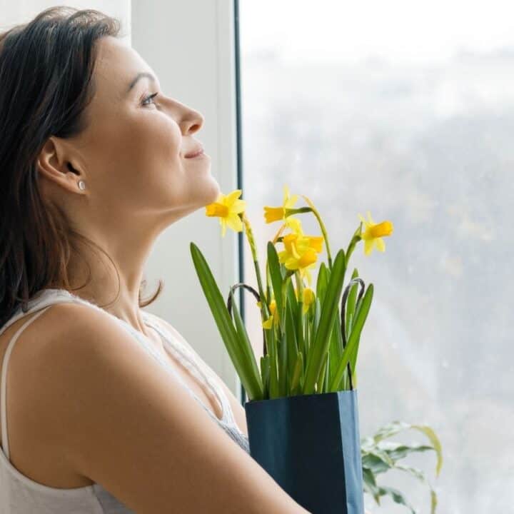 Woman holding a pot of yellow daffodils stands by a window, looking outside with a slight smile.