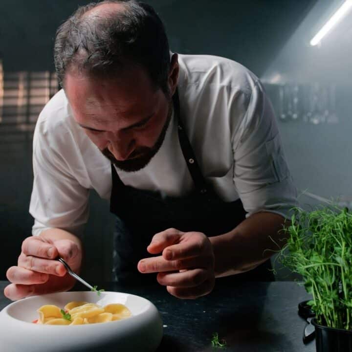 A chef in a dimly lit kitchen carefully garnishes a dish in a white bowl, with fresh herbs on the counter nearby.