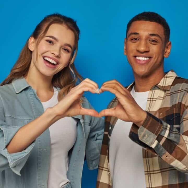 Two people standing close together in front of a blue background, smiling and forming a heart shape with their hands.