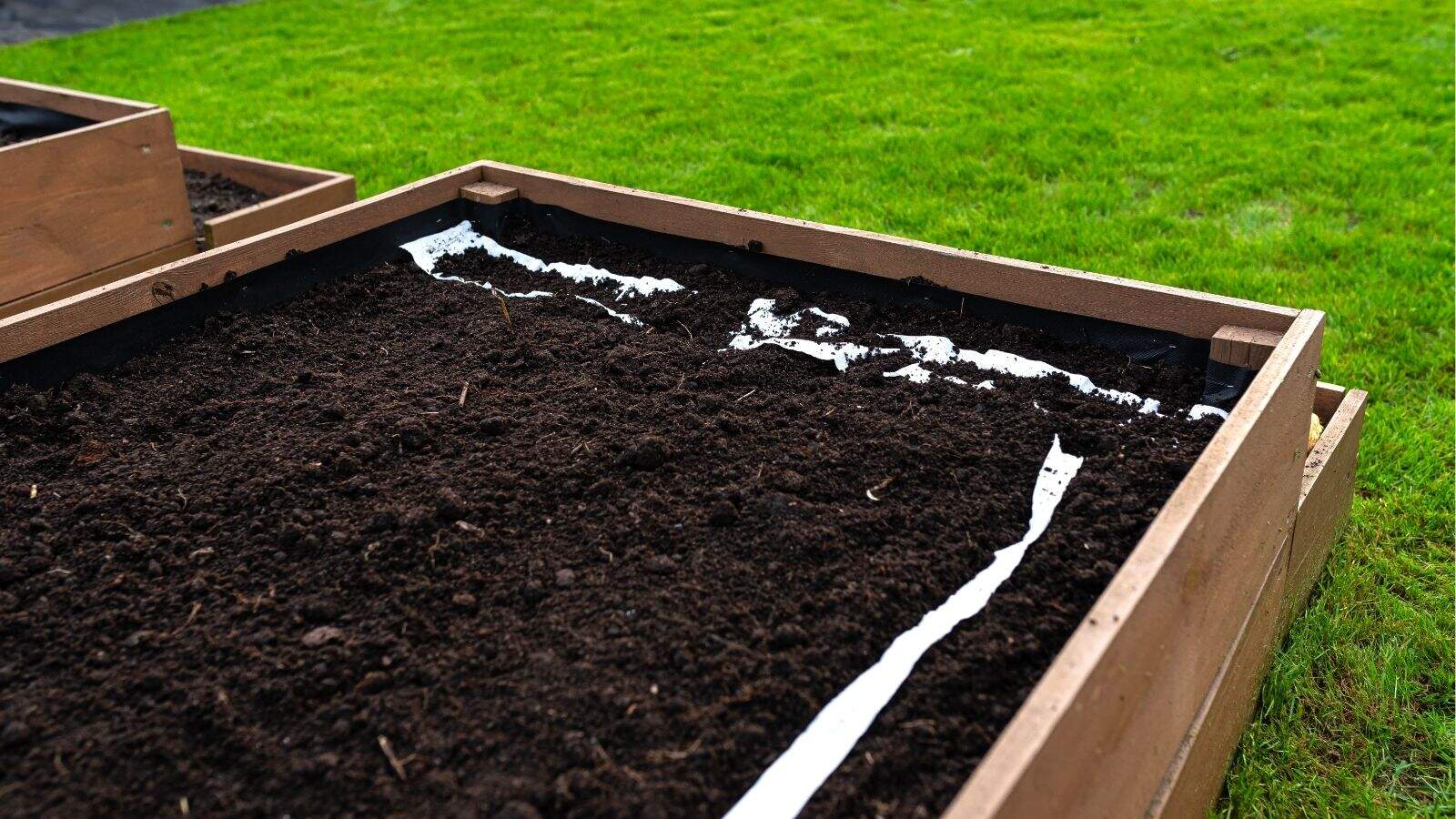 A wooden raised garden bed filled with dark soil stands on green grass, with white liner visible along one edge inside the bed.