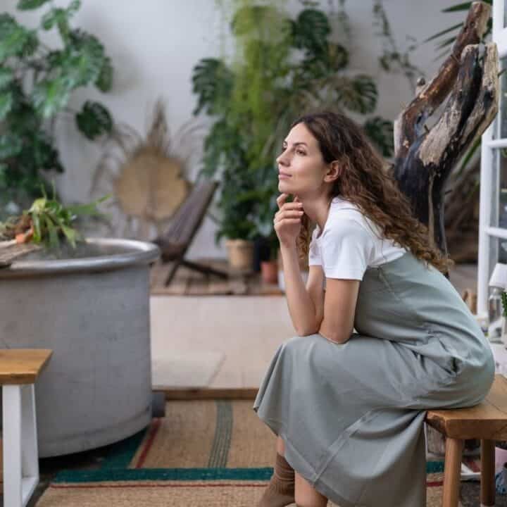 A woman sits on a wooden bench in a plant-filled room, resting her chin on her hand and looking thoughtful.