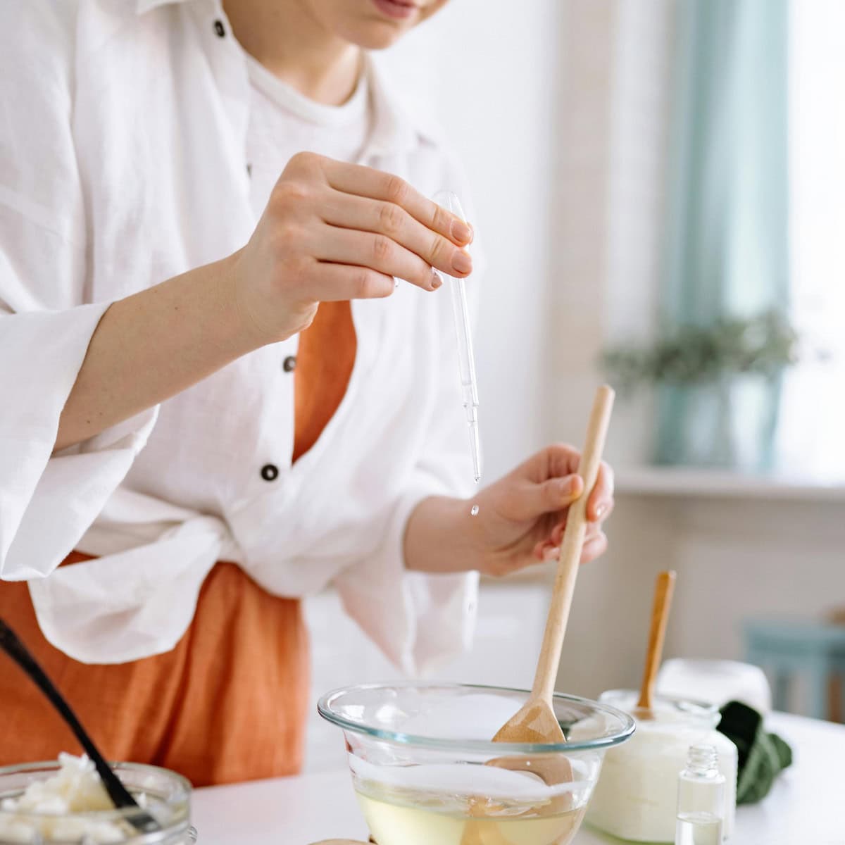 Person using a dropper to add liquid to a bowl while stirring with a wooden spoon in a bright kitchen.