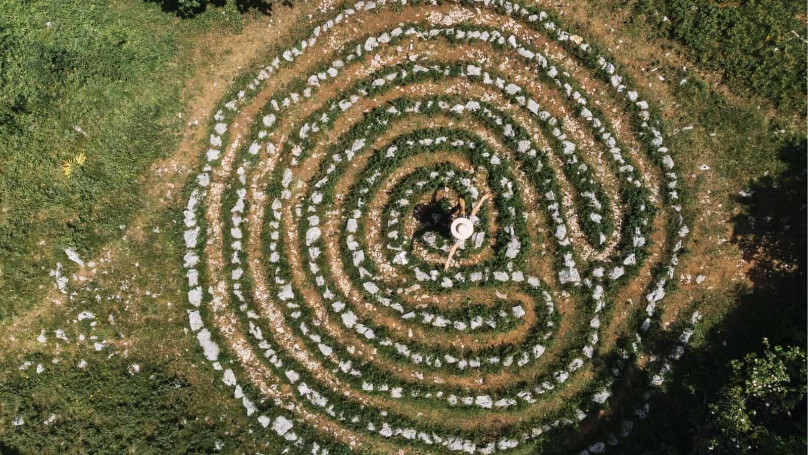 A person wearing a hat stands at the center of a circular stone labyrinth surrounded by grass, seen from above.