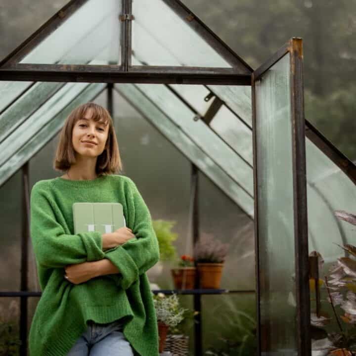 A person in a green sweater stands in the doorway of a greenhouse, holding a book and smiling, with plants visible inside and outside the structure.