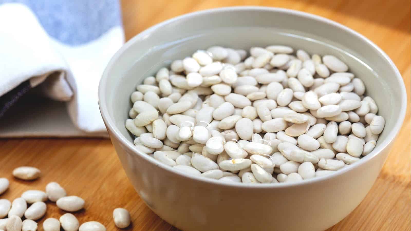 A bowl filled with white beans soaking in water sits on a wooden surface with some beans scattered nearby.