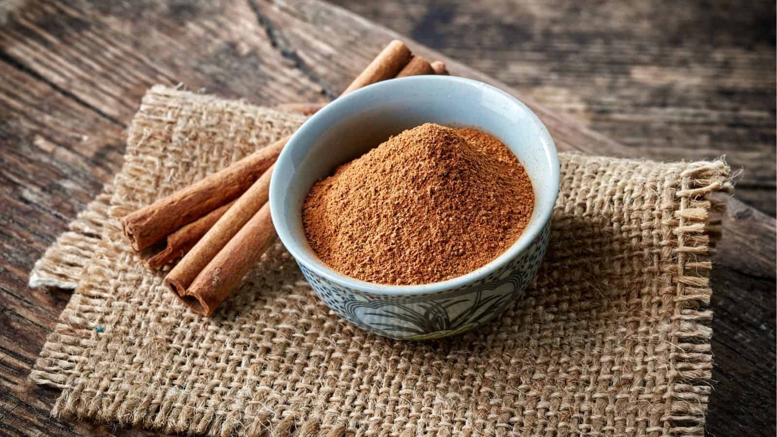 A bowl of ground cinnamon and cinnamon sticks placed on a piece of burlap on a wooden surface.