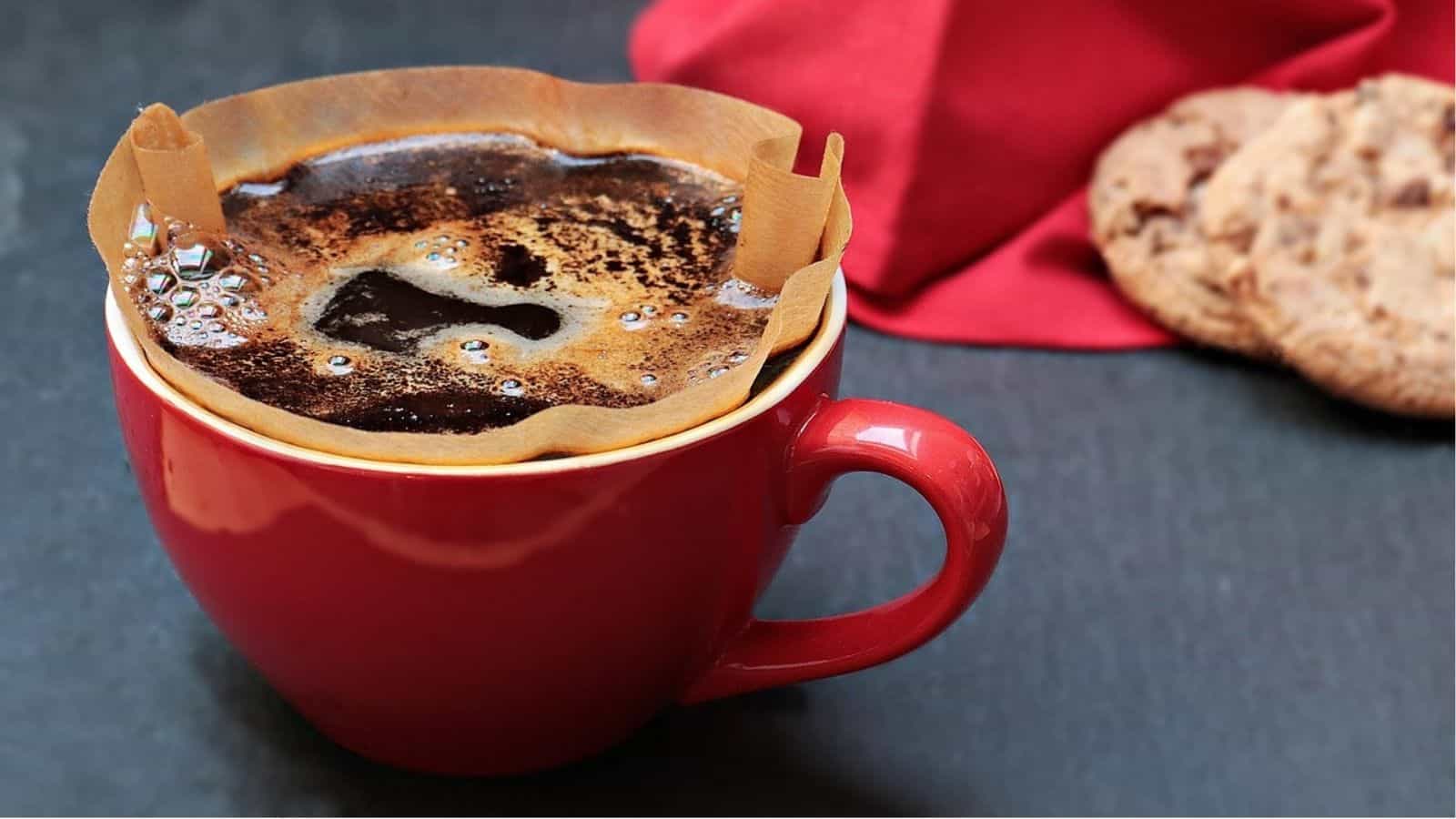A red mug with a coffee filter filled with brewed coffee sits on a dark surface, with cookies and a red napkin in the background.