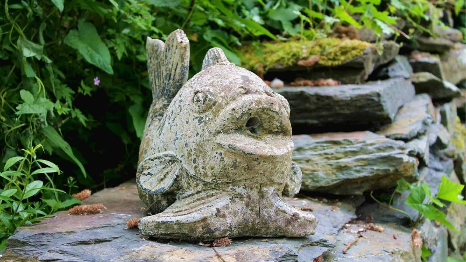 A weathered stone fish sculpture sits on layered rocks, surrounded by green plants and moss.