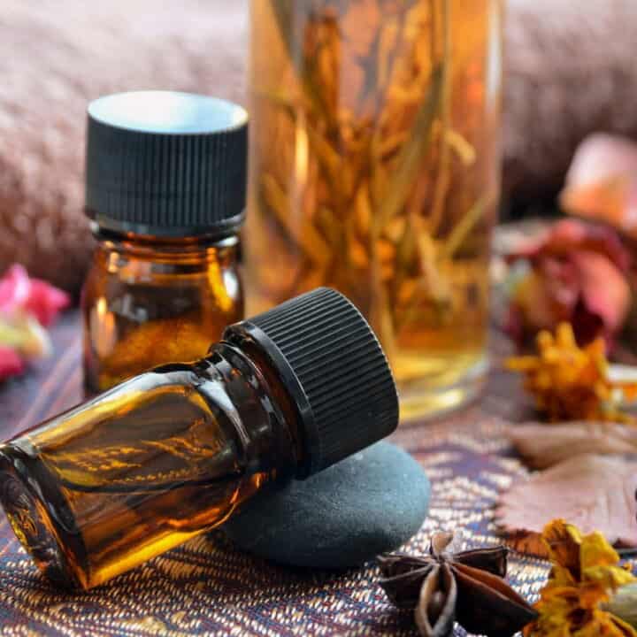 Amber glass bottles with black caps, one upright and one lying down on a stone, surrounded by dried flowers and a large jar of botanicals—perfect for those interested in cooking with essential oils.