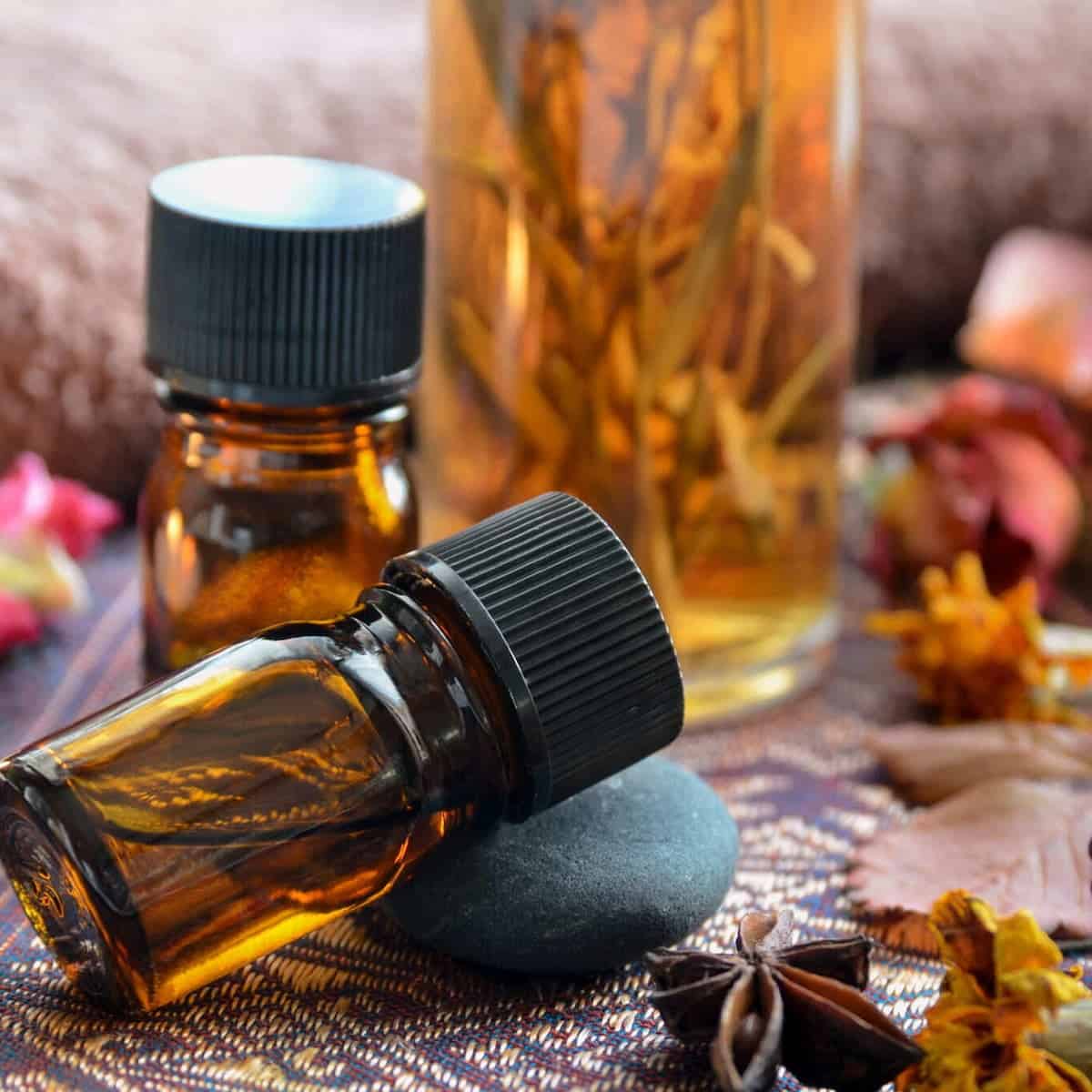 Amber glass bottles with black caps, one upright and one lying down on a stone, surrounded by dried flowers and a large jar of botanicals—perfect for those interested in cooking with essential oils.