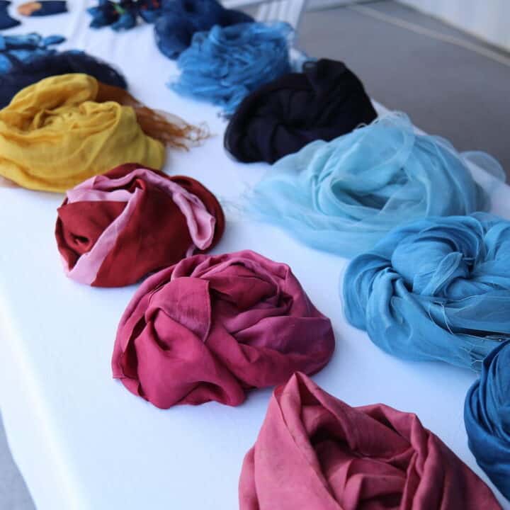 Scarves in various colors, including red, blue, yellow, and black, are neatly laid out in rows on a white tablecloth.