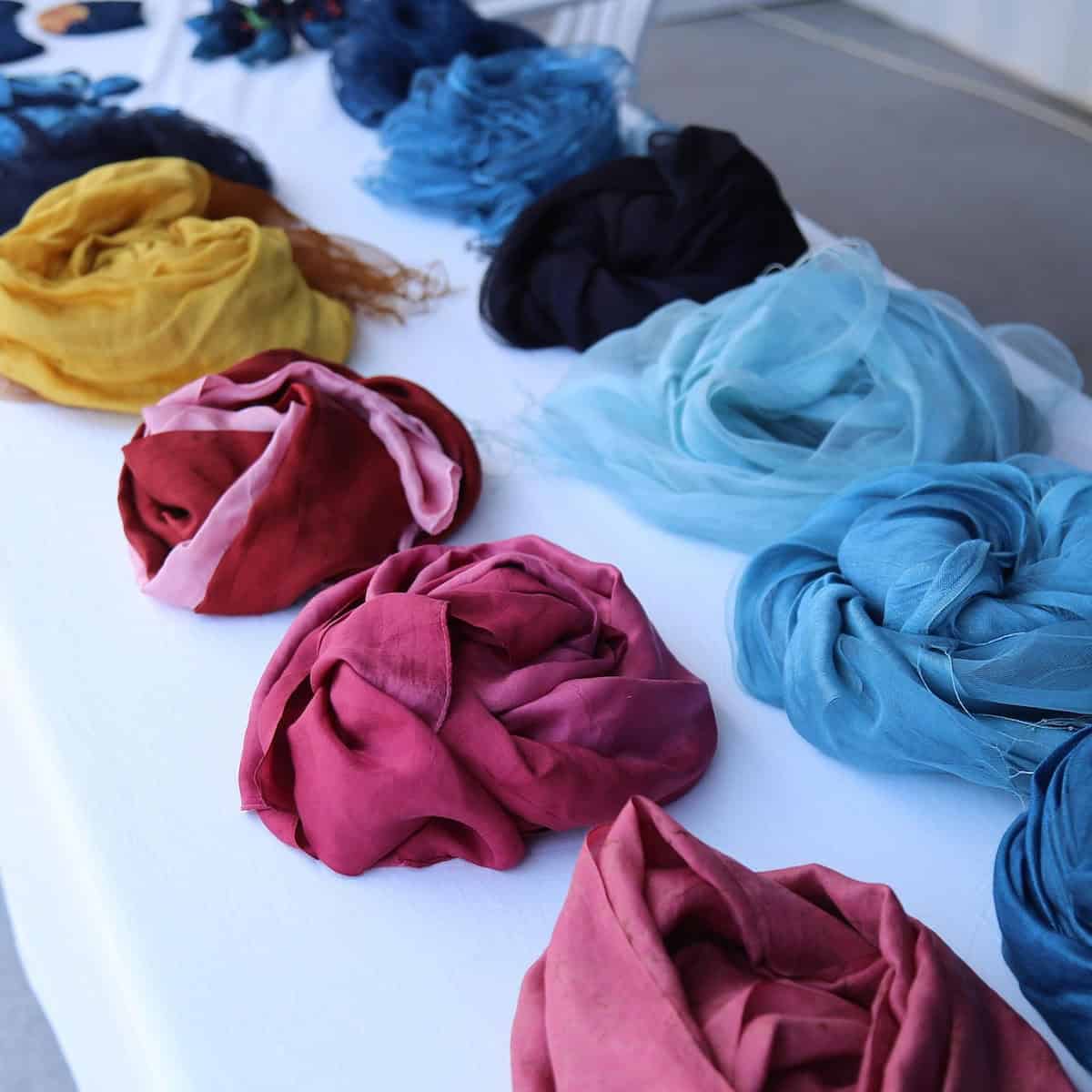 Scarves in various colors, including red, blue, yellow, and black, are neatly laid out in rows on a white tablecloth.