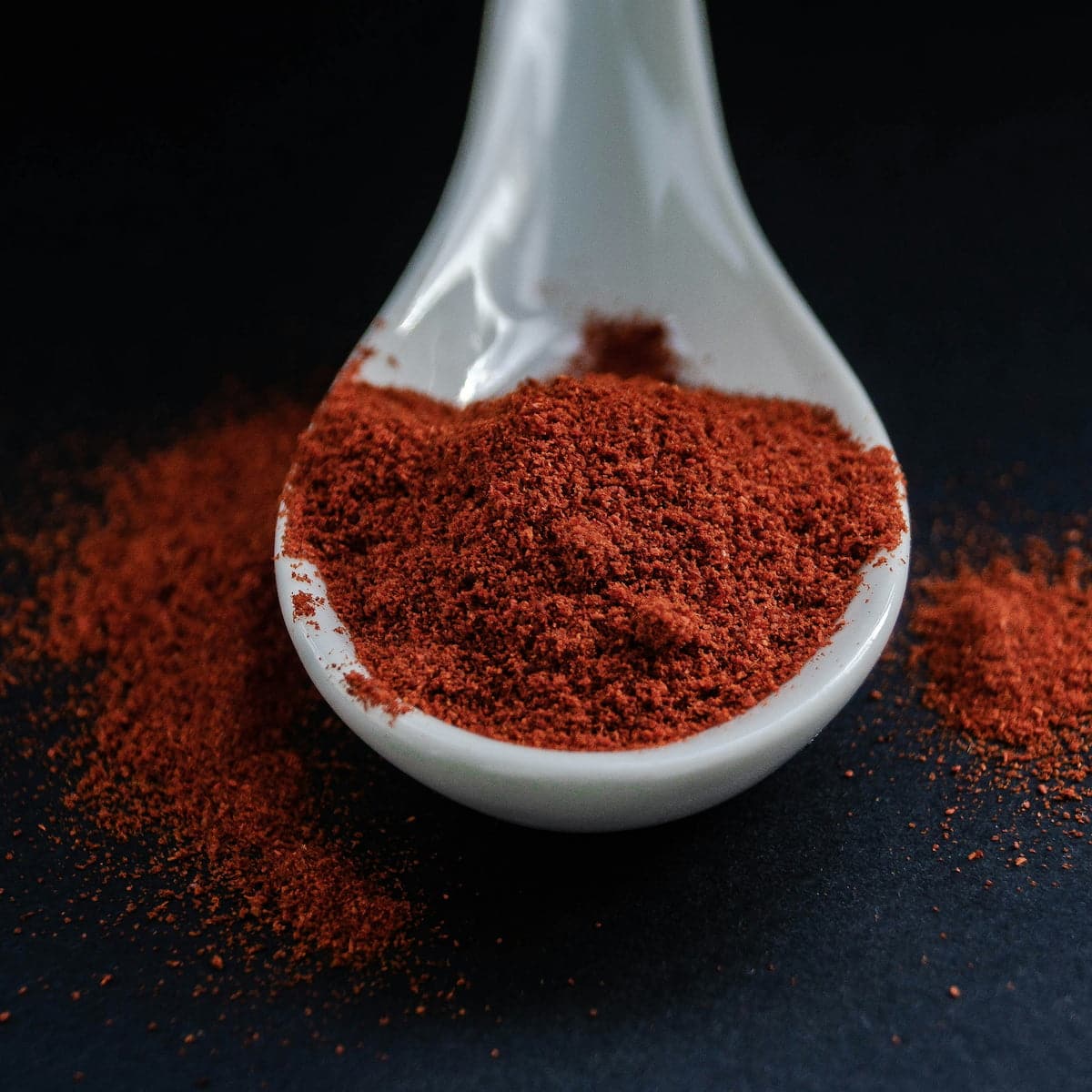 A white ceramic spoon holds a mound of finely ground reddish-brown spice, with more of the spice scattered on a dark surface.