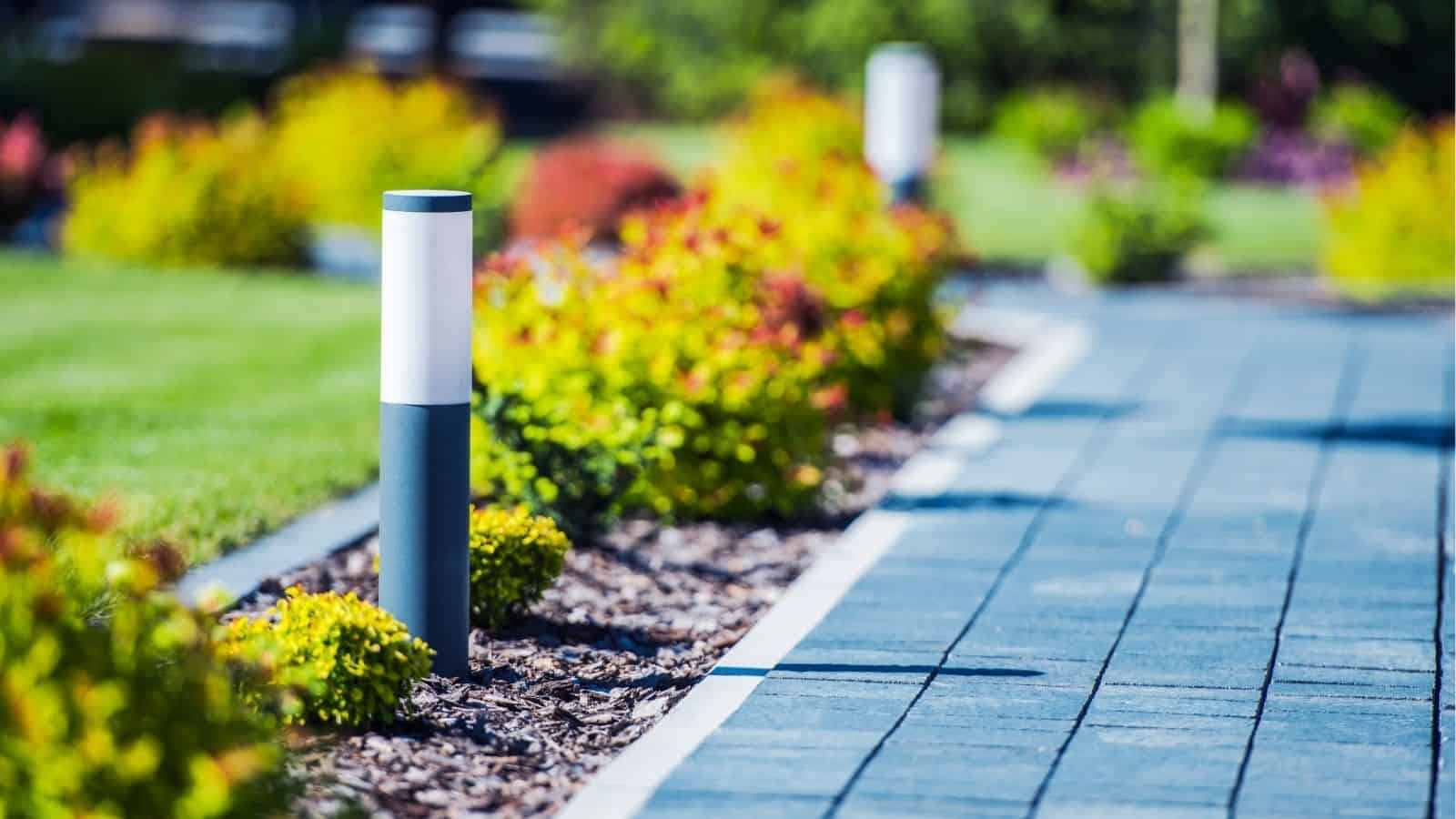 A modern outdoor pathway with gray pavers, lined with small garden lights and colorful shrubs, on a bright sunny day.