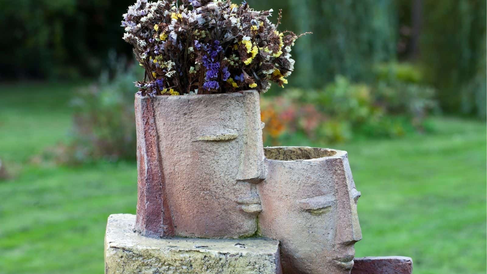 Two stone sculptures of abstract human faces, one with an open top containing dried flowers, are displayed outdoors on a grassy area.