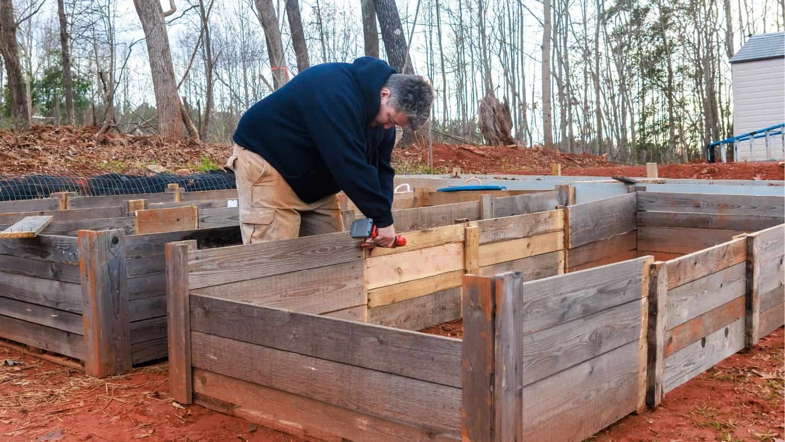A person uses a drill to assemble a wooden raised garden bed outdoors, surrounded by bare trees and soil.