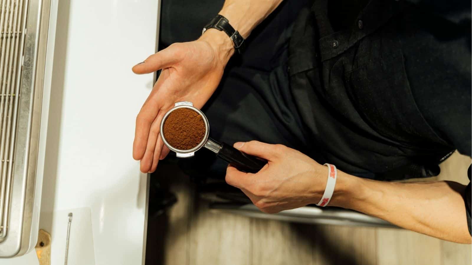 A person holding a portafilter filled with ground coffee, preparing for espresso by an espresso machine.