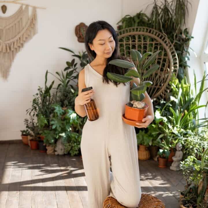 A woman in a beige jumpsuit holds a potted plant and a spray bottle, standing in a bright room filled with various indoor plants and natural decor.