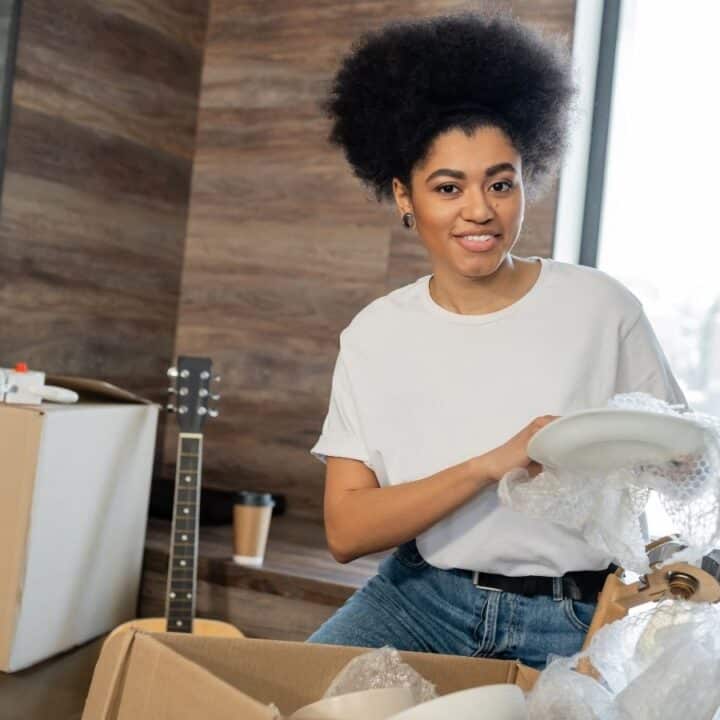 A woman in a white shirt wraps a plate in bubble wrap while packing or unpacking boxes in a bright room with a guitar and plants.