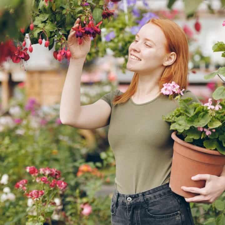 A woman with red hair holds a potted plant and examines hanging flowers in a garden or greenhouse.