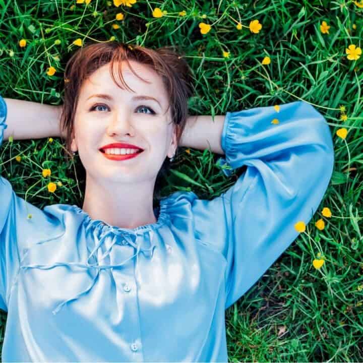A woman in a light blue blouse lies on green grass with yellow flowers, smiling and looking upward with her hands behind her head.