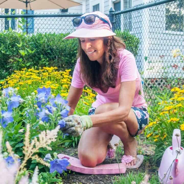 A woman wearing a pink hat and gloves tends to flowers in a colorful garden on a sunny day.