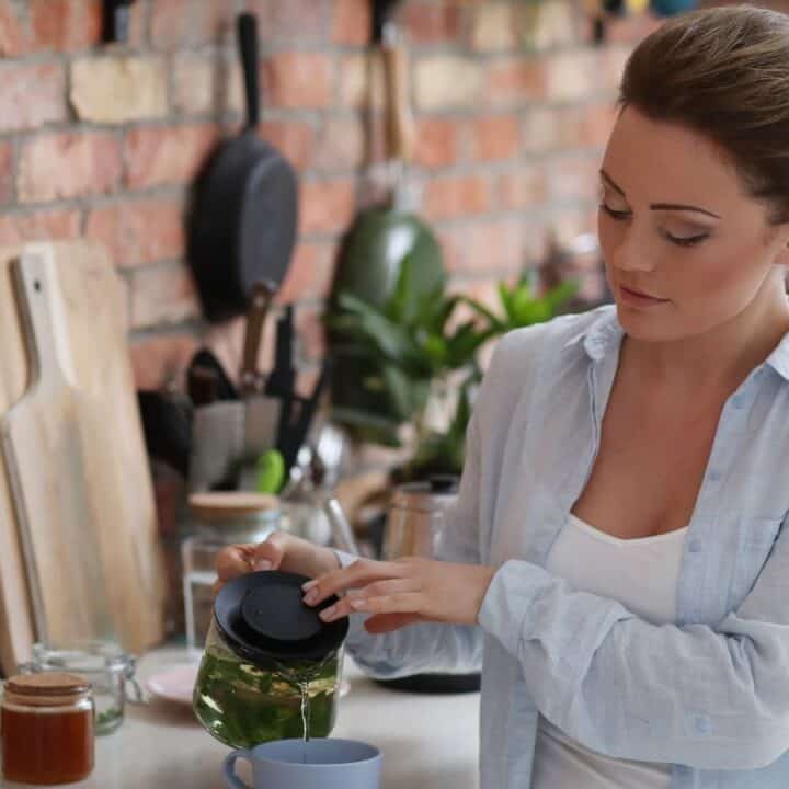 Woman in a kitchen pours tea from a glass teapot into a blue mug. A jar of honey, glass of water, and cutting boards are visible on the counter.