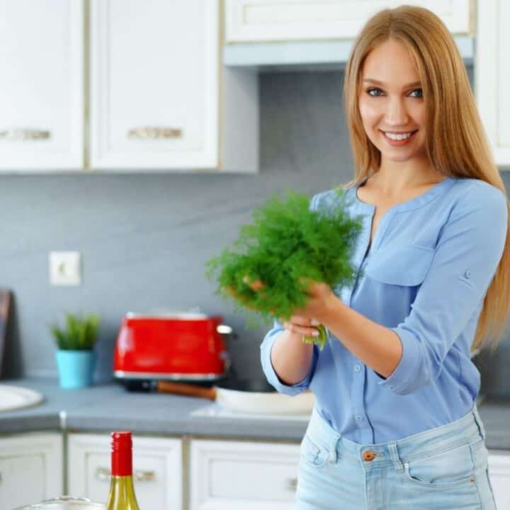 A woman in a blue shirt stands in a kitchen holding fresh green herbs, with a red toaster and bottles visible on the counter behind her.