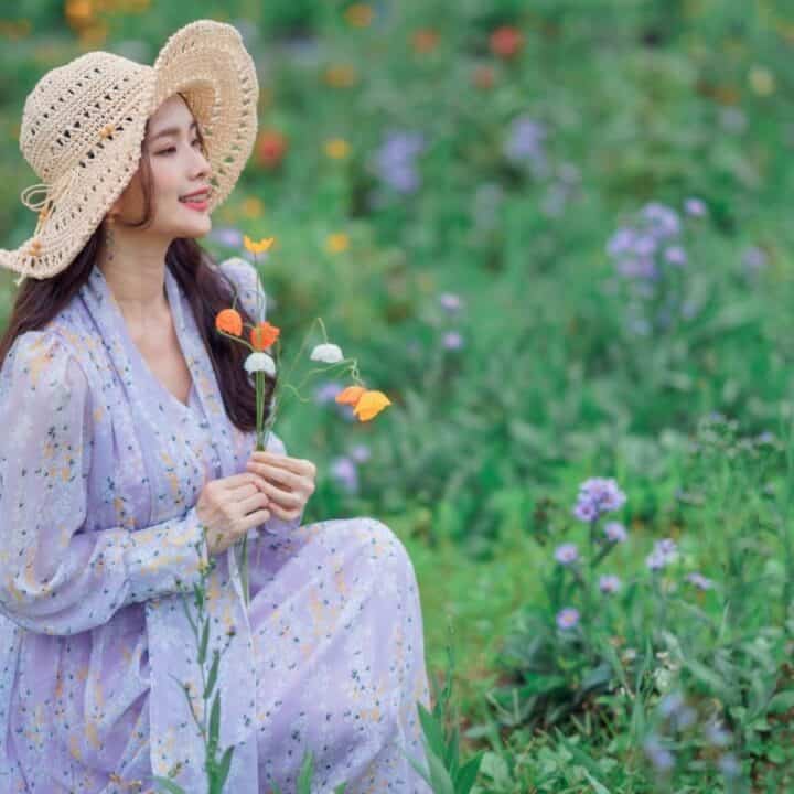 A woman in a light purple dress and straw hat sits in a field of wildflowers, holding orange flowers and looking ahead.